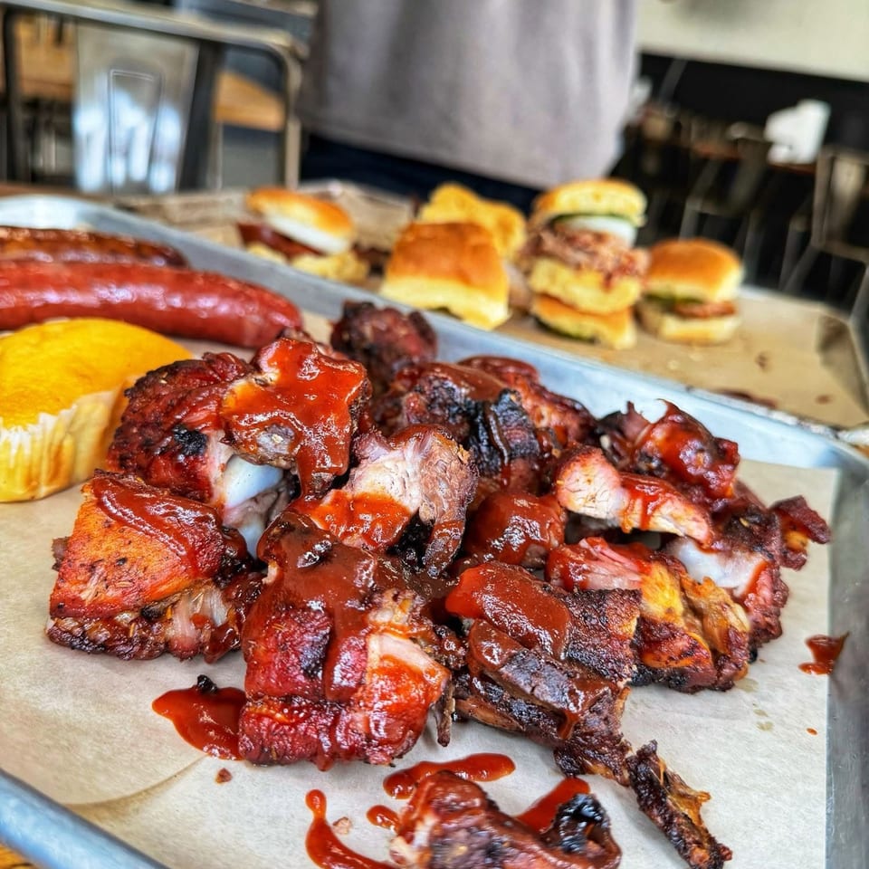 Close-up of barbecue ribs with sauce, sausage, cornbread, and sliders on a metal tray.