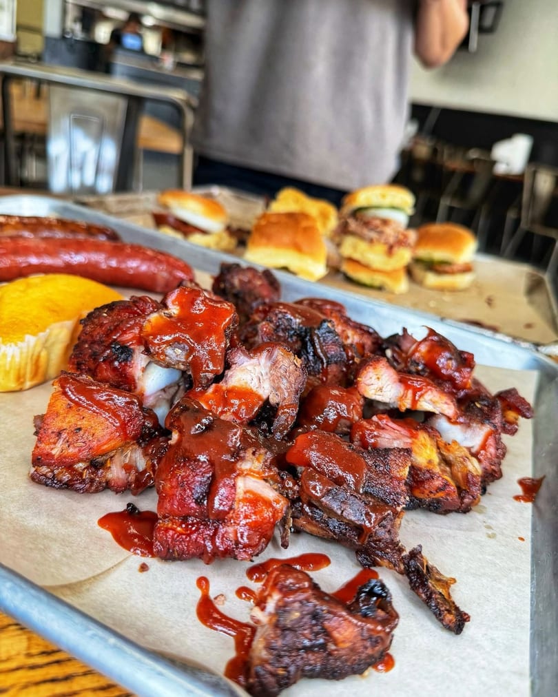 Close-up of barbecue ribs with sauce, sausage, cornbread, and sliders on a metal tray.