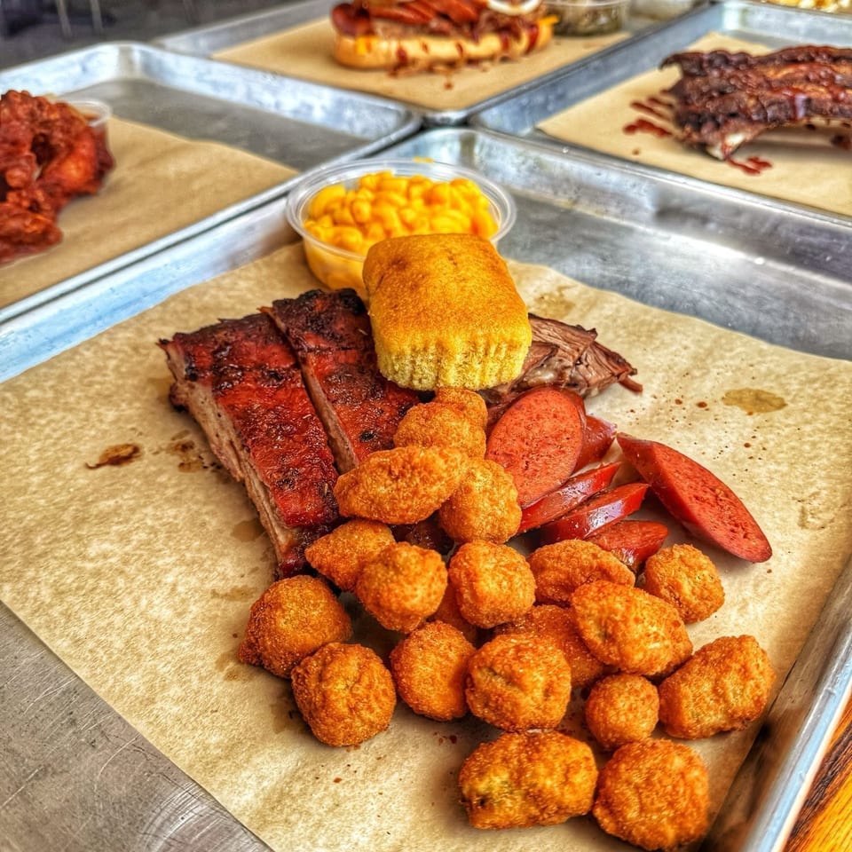 A BBQ platter with ribs, sausage, hush puppies, cornbread, and mac and cheese on a metal tray.