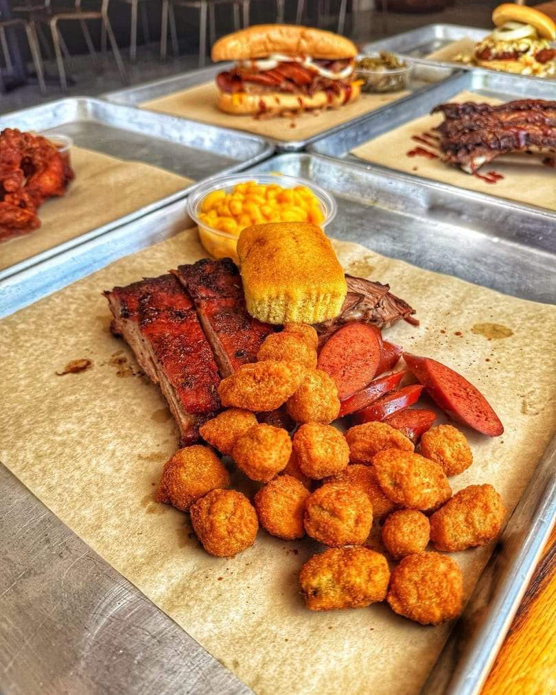 A BBQ platter with ribs, sausage, hush puppies, cornbread, and mac and cheese on a metal tray.