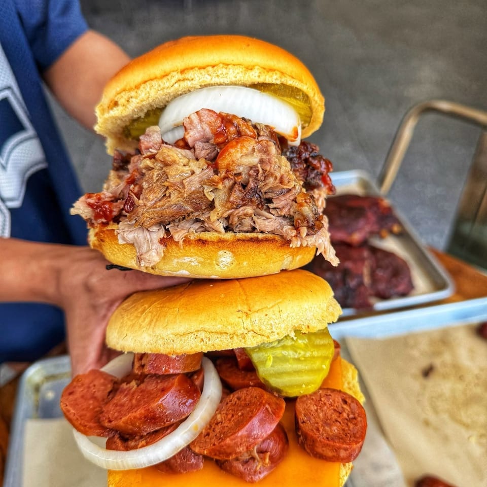 Two hands holding a stacked pulled pork sandwich and a sausage, cheese, and pickle sandwich.