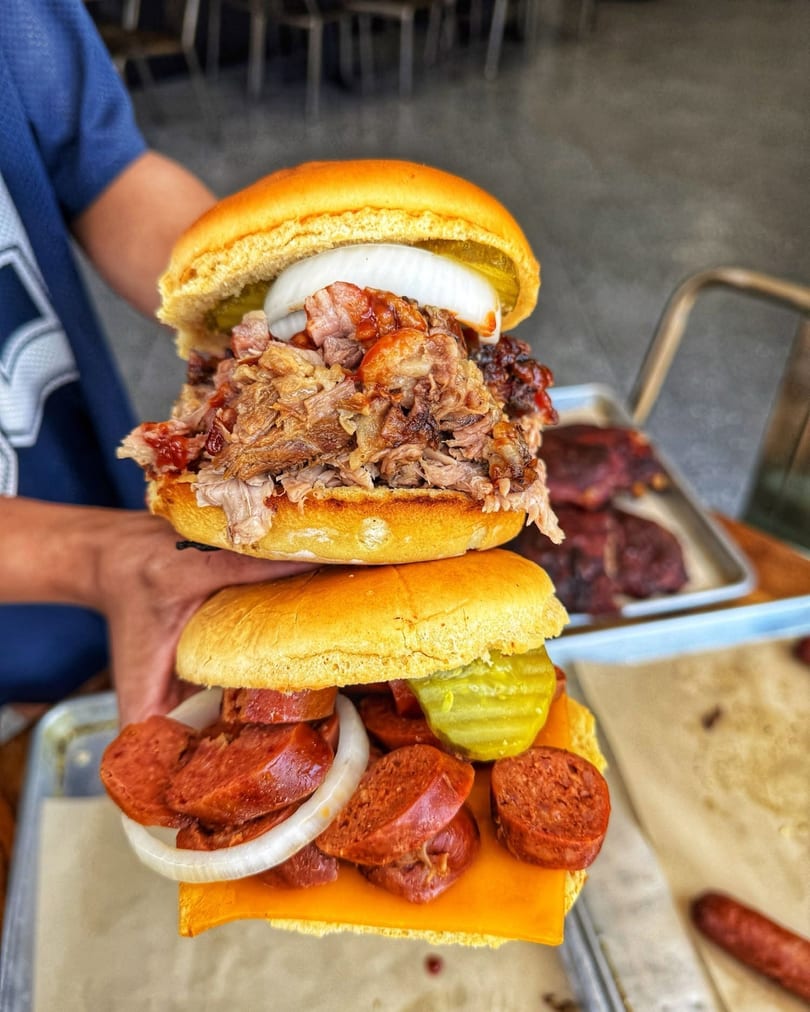 Two hands holding a stacked pulled pork sandwich and a sausage, cheese, and pickle sandwich.
