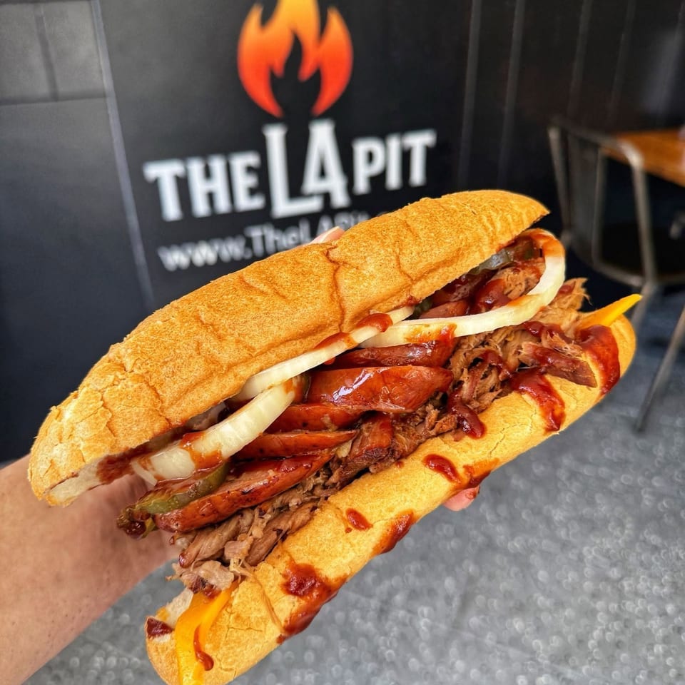 A large BBQ sandwich with pulled pork, sausage, onions, and pickles, held in front of The La Pit sign.