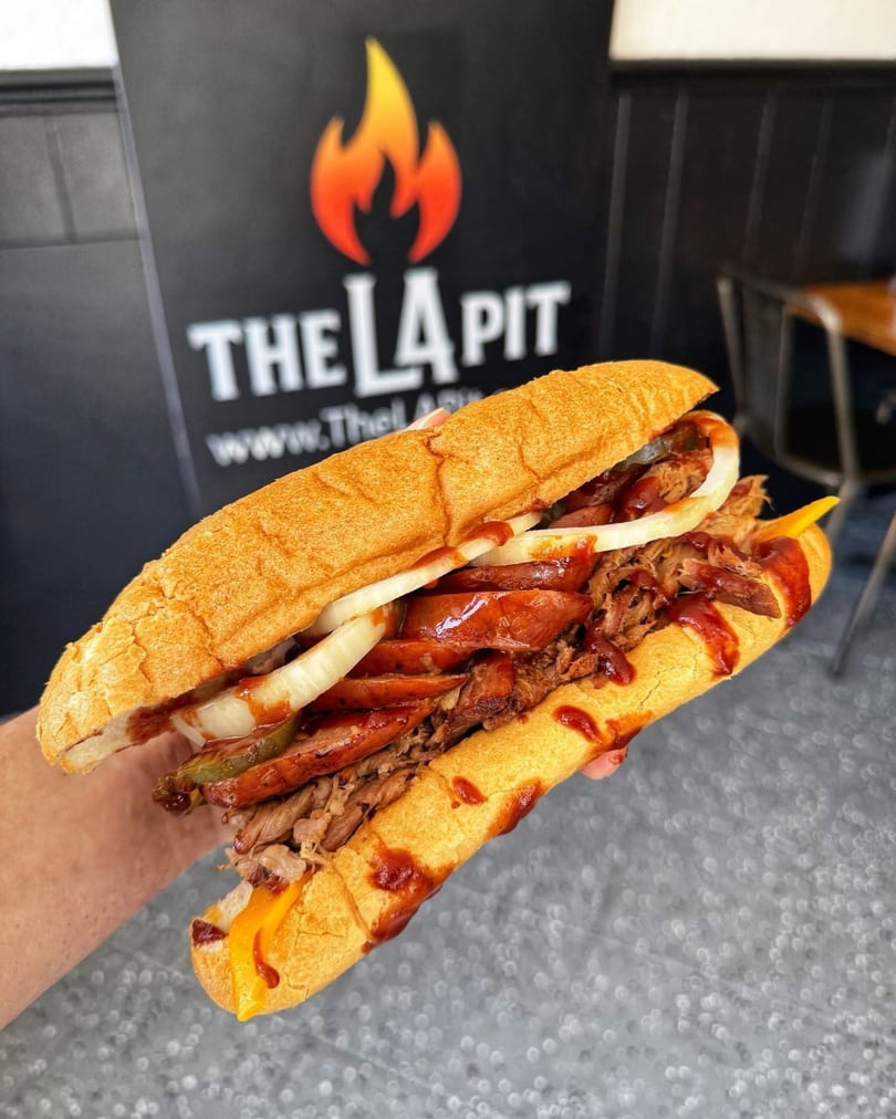 A large BBQ sandwich with pulled pork, sausage, onions, and pickles, held in front of The La Pit sign.