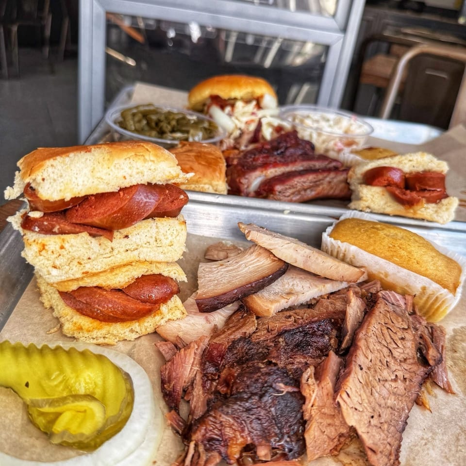A tray of barbecue food including brisket, ribs, sausage sandwiches, corn bread, and sides.