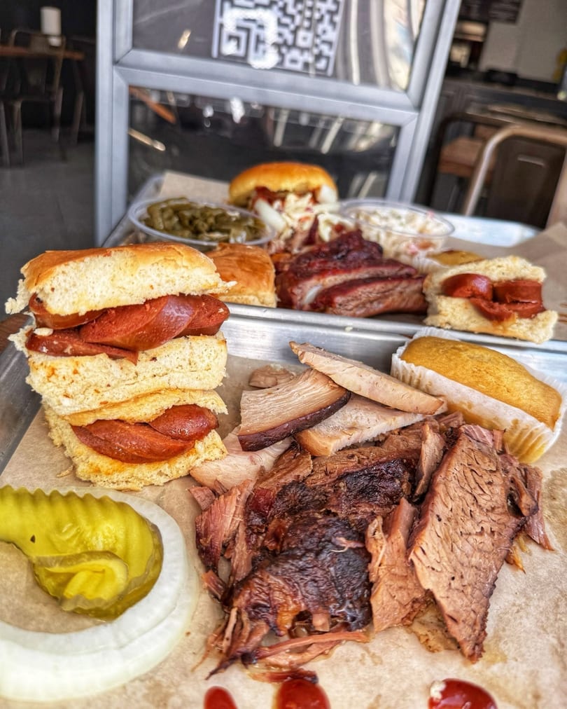 A tray of barbecue food including brisket, ribs, sausage sandwiches, corn bread, and sides.